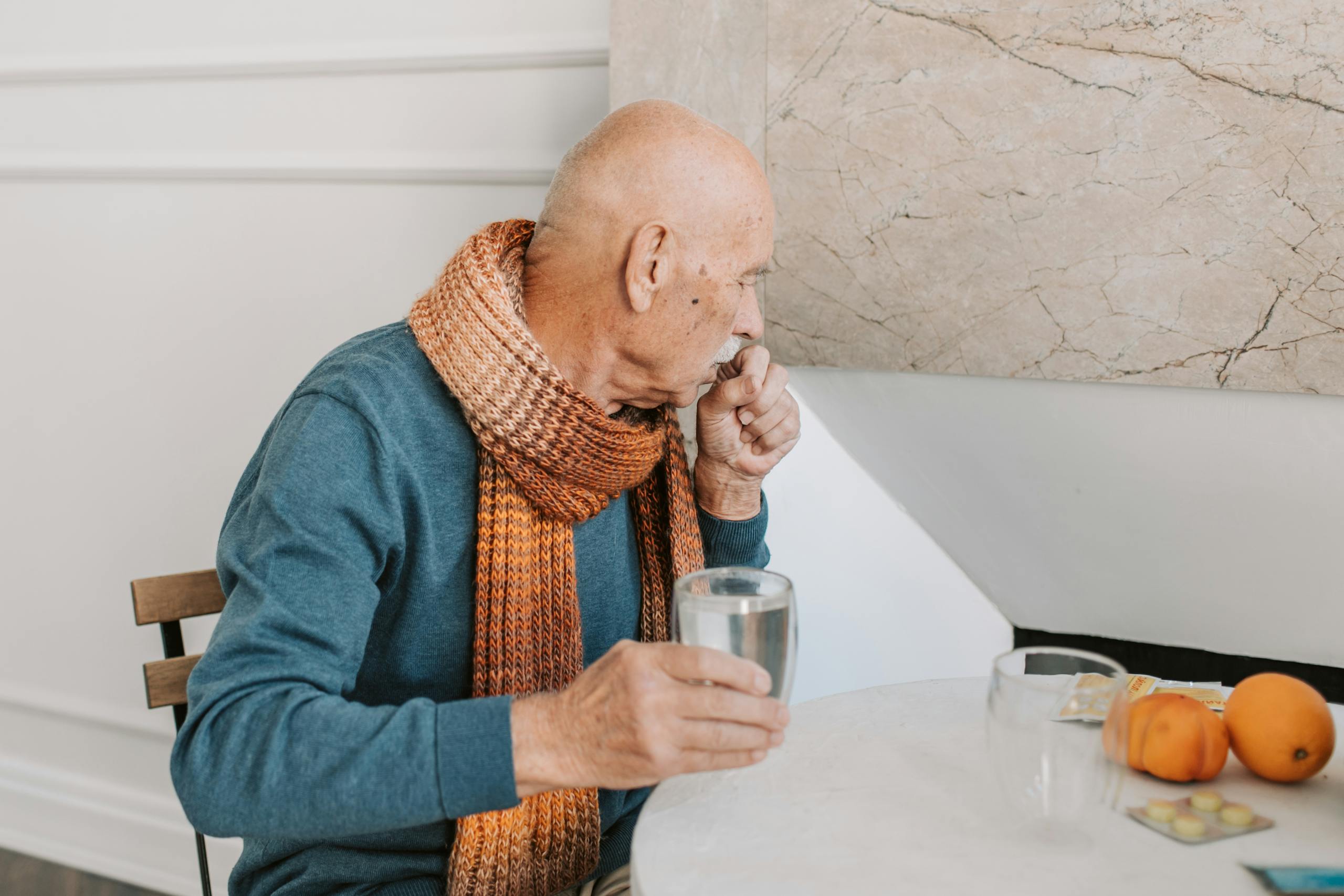 Elderly man in blue sweater coughing, holding water at white table with medicines.